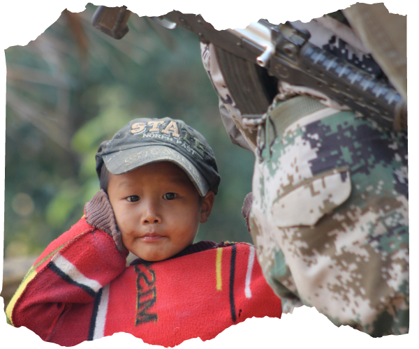 A young boy from Kachin State Burma is stood behind a solider with a gun. He has a baseball cap and a red jumper on. He is looking directly at the camera and has his hand over one ear. 