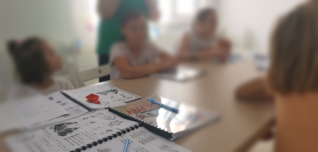 Focus on a pile of school books, with children around the table listening to an adult. Their faces are blurred. 