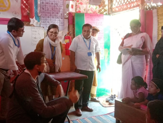Veena, a middle aged Indian woman wearing a white scarf is smiling as she visits a colourful classroom in Kutupalong camp with Bangladesh staff from Mukti