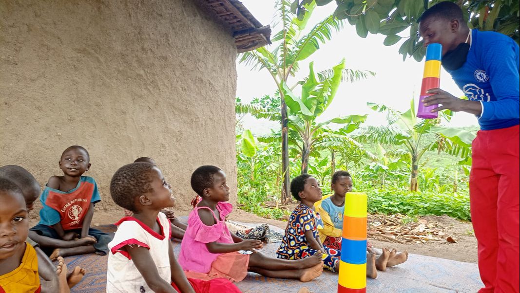 A group of young Congolese refugee children learning outside. They are sat on woven mats and are looking up at their teacher who is holding some coloured stacking cups.
