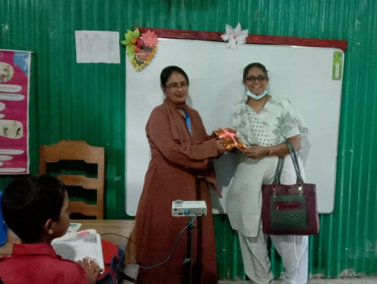 Veena accepting a gift from a teacher in a classroom with green corrugated walls and a blank whiteboard decorated with flowers in Bangladesh