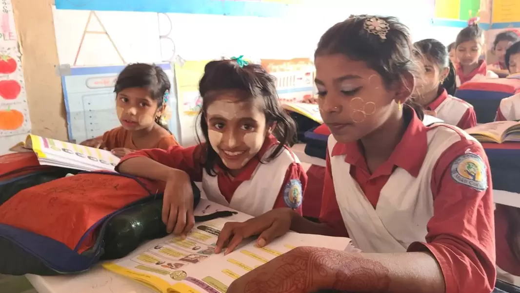 Three Rohingya girls wearing school uniform sat at their desk with workbooks and bags