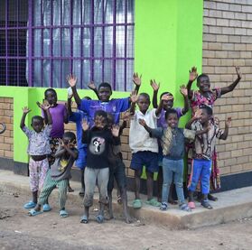 Young children are excited to be standing in front of their brand new classroom in Kyaka II. They are standing in front of a new brick building with bright green walls and purple grated windows.