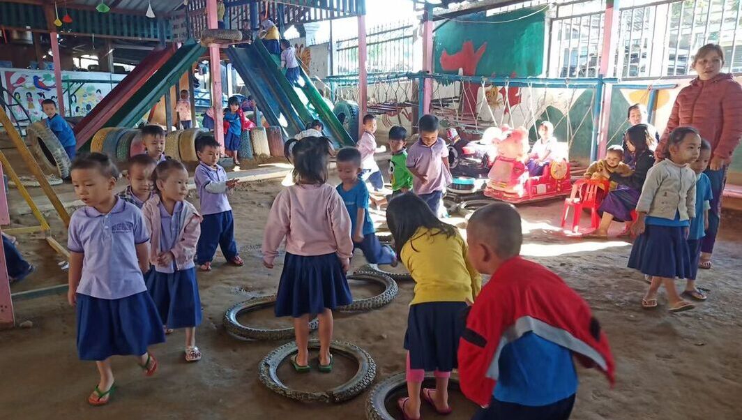 Kachin children playing on bright play equipment in the sandy covered garden of their preschool centre in Myanmar