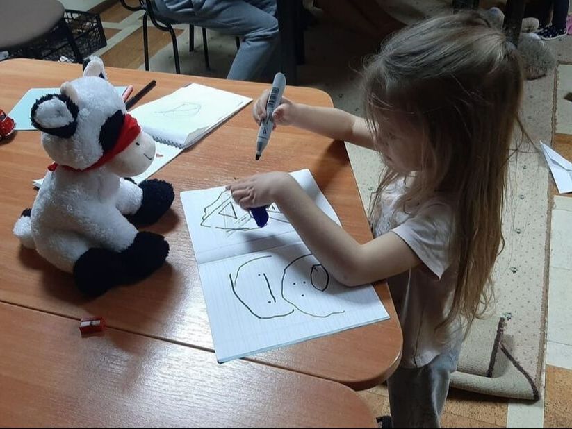 Young Ukrainian refugee girl drawing a stuffed toy sheep at a table