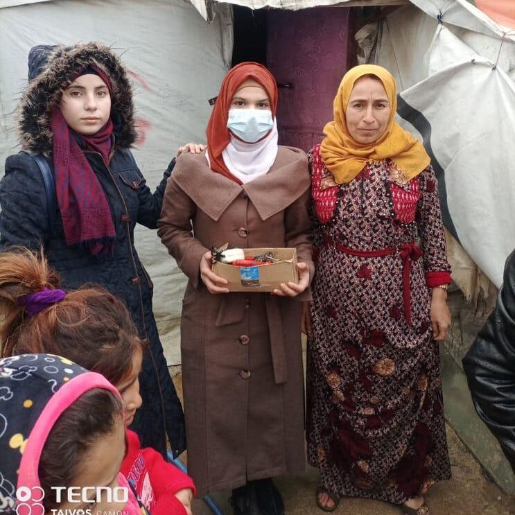 Grade 9 student standing with two women in headscarves outside a tented home. The women have just been given a small box of clothes.