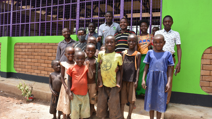 A group of Congolese refugee children and adults stand outside the bright green and purple new school building in Kyaka II, smiling.