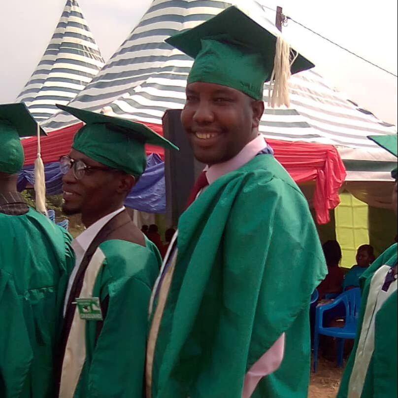 Muhorana John a young man from the DRC is wearing a green graduation cap and gown at an outside ceremony in Uganda. He smiling and standing with his fellow graduates.
