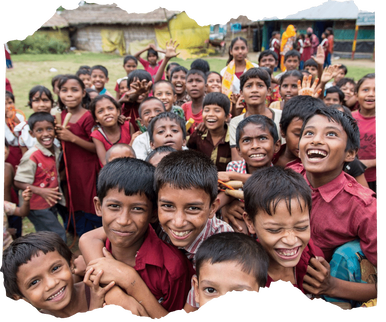 A large group of Bangladeshi children outside their community school in Cox's Bazar, Bangladesh. They are crowded around the camera, looking at it and smiling. Most of the children are wearing red school uniforms. 