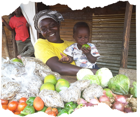 A Ugandan woman in a bright yellow T shirt sits behind her vegetable stall with her child on her knee. She is smiling at the camera and he is eating a piece of fruit