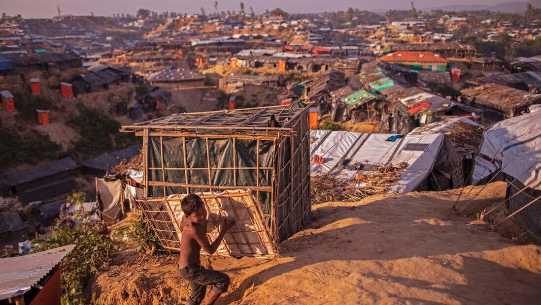 Scene of Kutupalong mega camp shortly after the August 2017 genocide. Photo shows miles of hastily constructed tents of sticks, mud and plastic and a small boy carrying a a slatted piece of wall. Picture