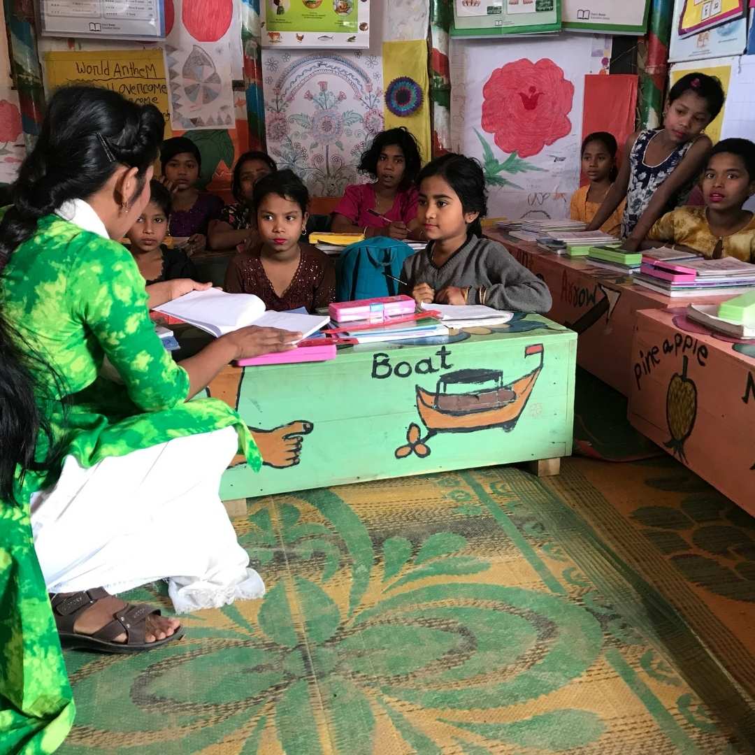 Teacher and class in Kutupalong refugee camp. Teacher is crouching down and chatting with girls who are sat at a colourful painted desk with a picture of a boat on it. 