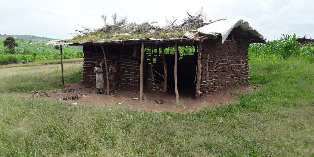 Image shows a dilapidated mud hut in a amongst a small area of grass. The hut is in much need of repair and has holes in the walls and roof. There are two young Congolese refugee children standing outside it. The hut is an existing early years classroom in Kyaka II settlement.