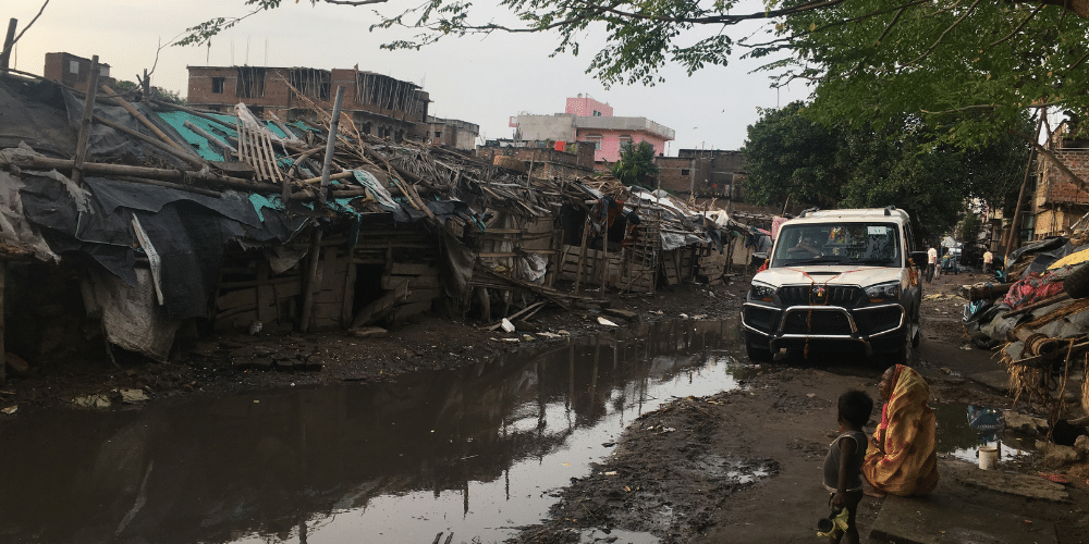 A flooded slum street in Bihar State with a car driving through. Makeshift shelters line the street and a small boy and older woman are sat outside on shelter. 