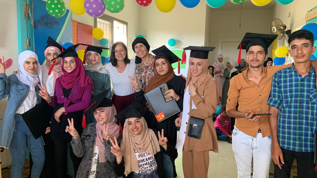 A group of Grade 9 Syrian students are stood inside their school surrounded by colourful balloons. They are wearing graduation hats and smiling. 