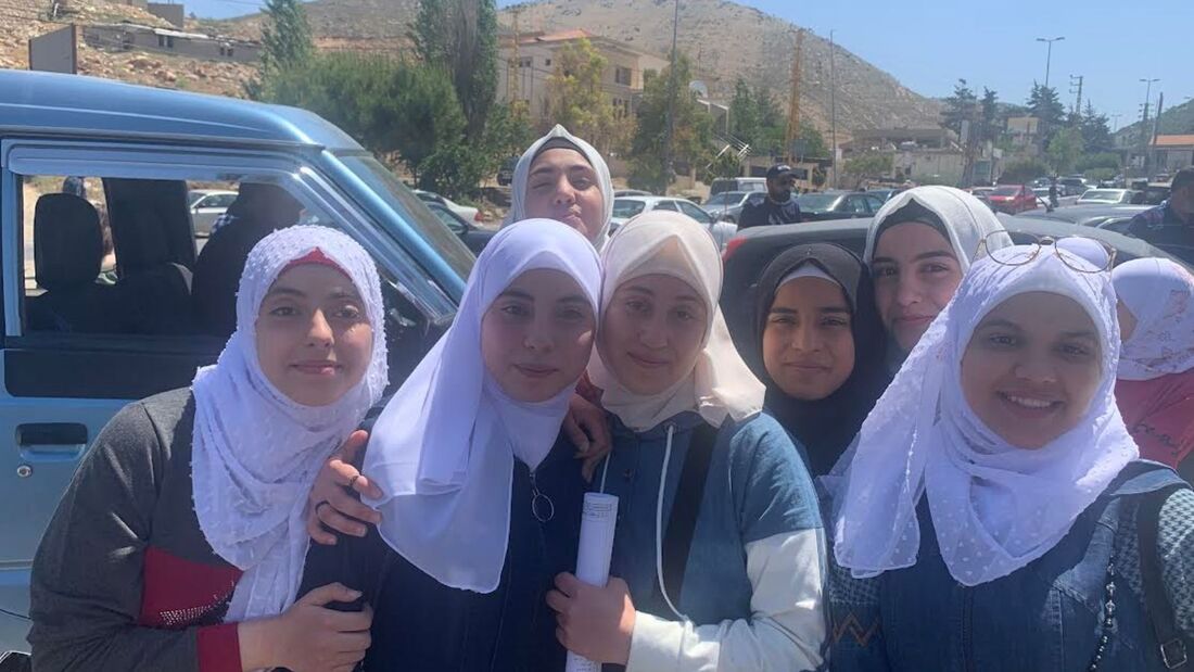 A group of teenage students, wearing headscarves are gathered together outside a minibus smiling at the camera before they set off to cross the border from Lebanon into Syria