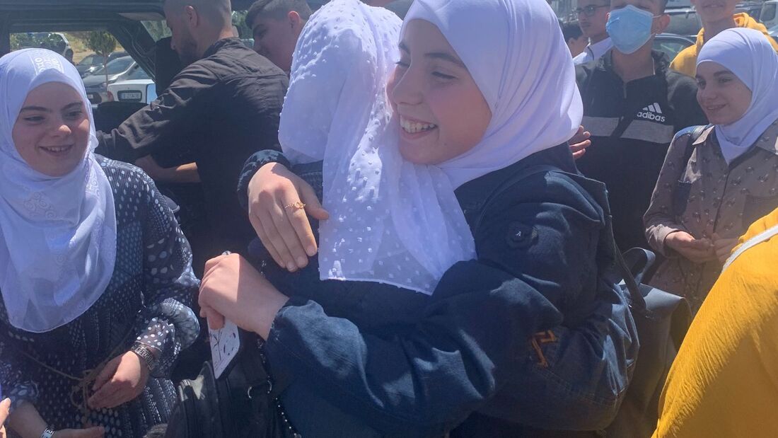 A Syrian student is wearing a white headscarf and smiling as she hugs someone