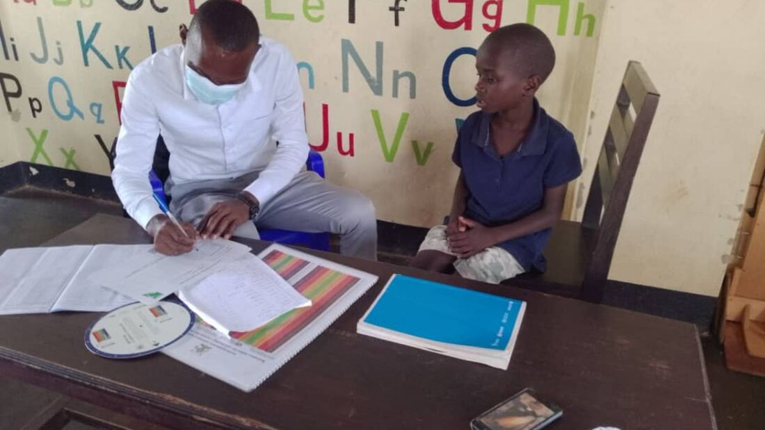 Healthcare professional wearing a wight shirt and mask writing notes on a piece of paper at a table with a young Ugandan boy in a blue tshirt and beige shorts