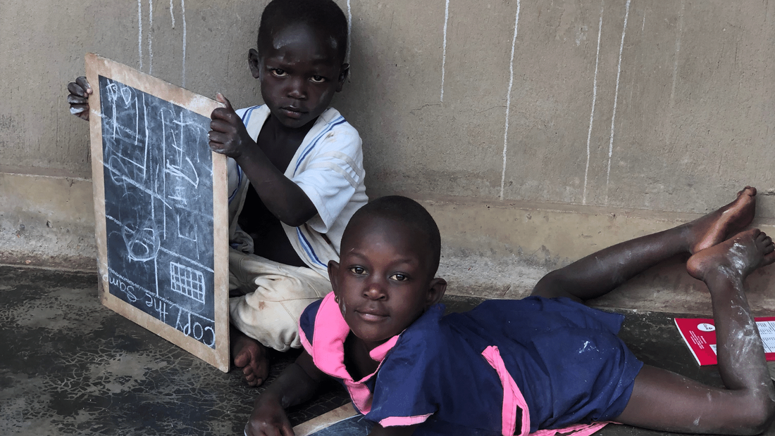 Two Ugandan children sat outside their home learning with slate boards and chalk.