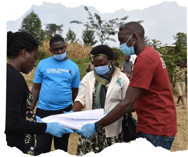 Image shows the team from Children on the Edge Africa. Four team members, two women and two men are standing outside holding a large piece of paper which they are all looking down at. Three of the team are wearing facemasks and blue rubber gloves. One man is wearing a blue tshirt with the children on the edge logo on the front. 