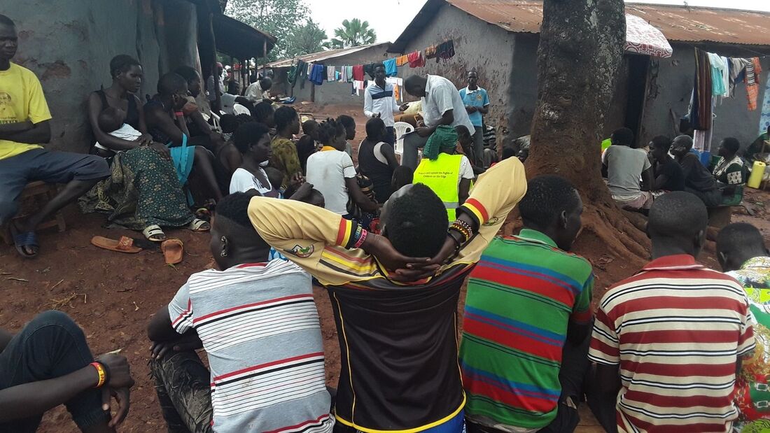 A group of Uganda parents are gathered around a tree in their slum community to listen to a workshop. They are sat around on chairs and the floor listening to the workshop leader.
