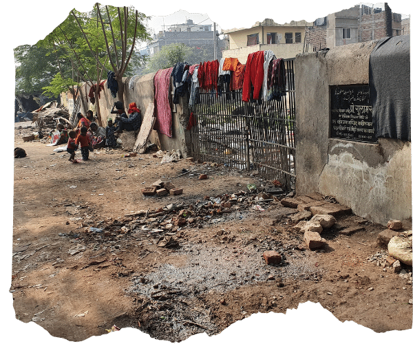 A dirty street lined with rubble and muddy puddles depicting typical conditions for a slum community in urban areas of Patna, India. Children are playing and adults sat along the wall lining the street.