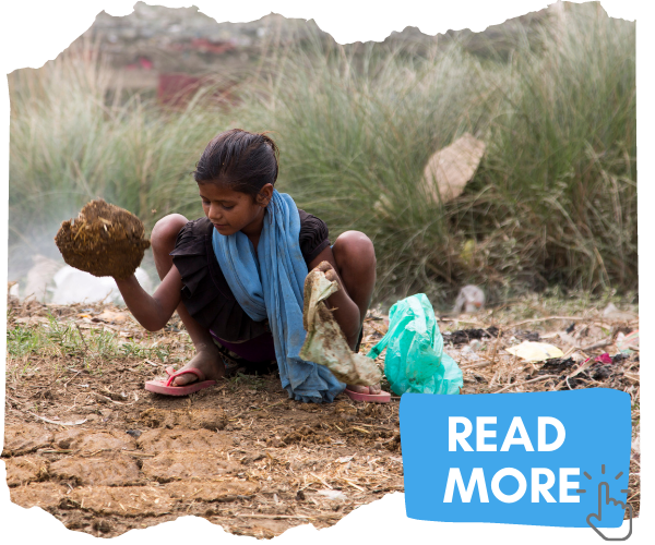 A young Dalit girl crouching on the muddy ground in front of some long grass. She is holding a handful of something brown and has a metal bowl in front of her on the ground. She is wearing a blue scarf and black dress with red flip flops. Litter is on the ground around her. Click on the image to read more about the issues in India