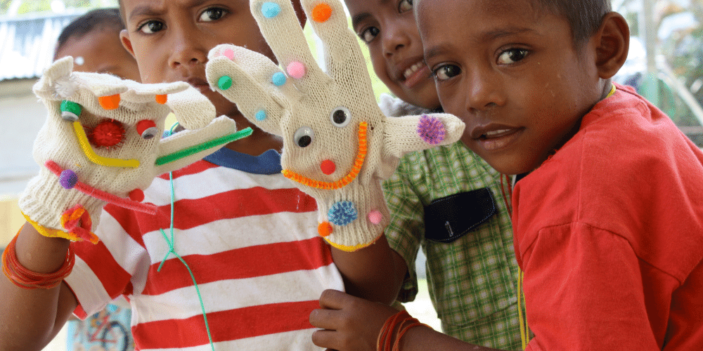 Three young Indonesian boys are looking at the camera with inquisitive looks on their faces. They are wearing bright clothes and two of the boys are holding puppets made of white gloves decorated in pom poms, googly eyes and pipe cleaners.