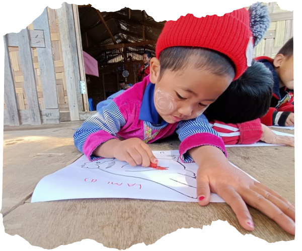Boy from Kachin State, Myanmar (about five years old) is lying on his front doing some colouring. Some of his classmates are doing the same behind him. He is wearing a bright jumper and red wooly hat as they are outside on some decking. He is slightly smiling as he works. 