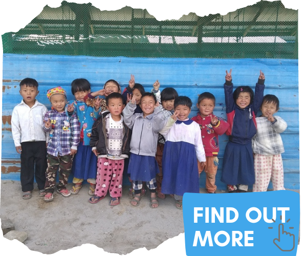 A group of 12 young children from Kachin State Myanmar (around 4 years old) are posing for a photograph in front of a blue building. They are smiling and some are waving or have their hands in the air. You can click this image to be taken to a page to find out more. 