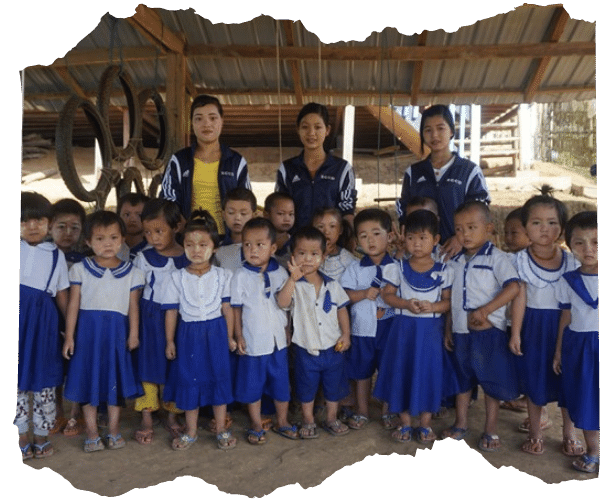 Image shows three Kachin women standing behind a large group of preschool children from an ECD centre in Kachin State, Myanmar. The children are all wearing blue and white school uniform and are looking at the camera. The three women at the back are wearing blue sports jackets and are also looking at the camera. They are all stood undercover in an open-sided building with a tin rood and play equipment visible behind them. 