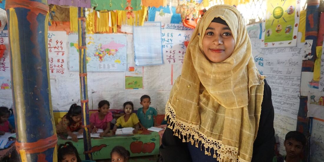 Image shows a young female teacher wearing a yellow headscarf and smiling. Behind her is a colourful classroom with lots of artwork on the walls, painted desks and children learning. 