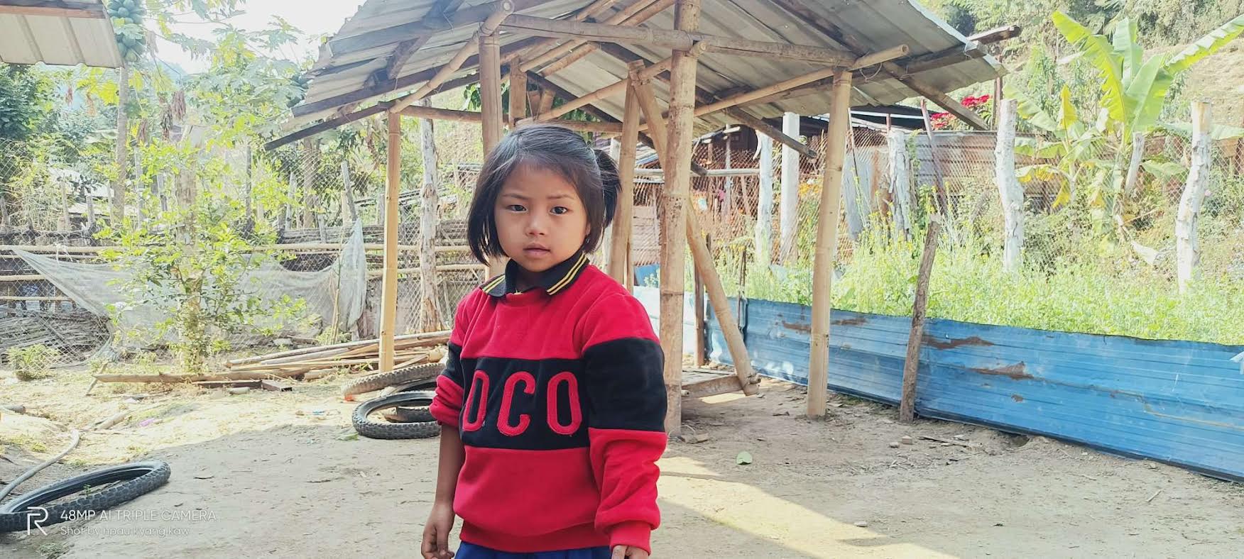 Kawn Ja Htoi Tsin is wearing a red and black jumper stood outside in a garden in Kachin State