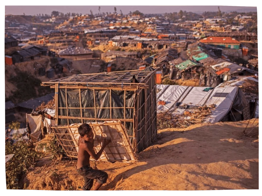 A Rohingya boy carrying bamboo against the backdrop of a sprawling refugee camp
