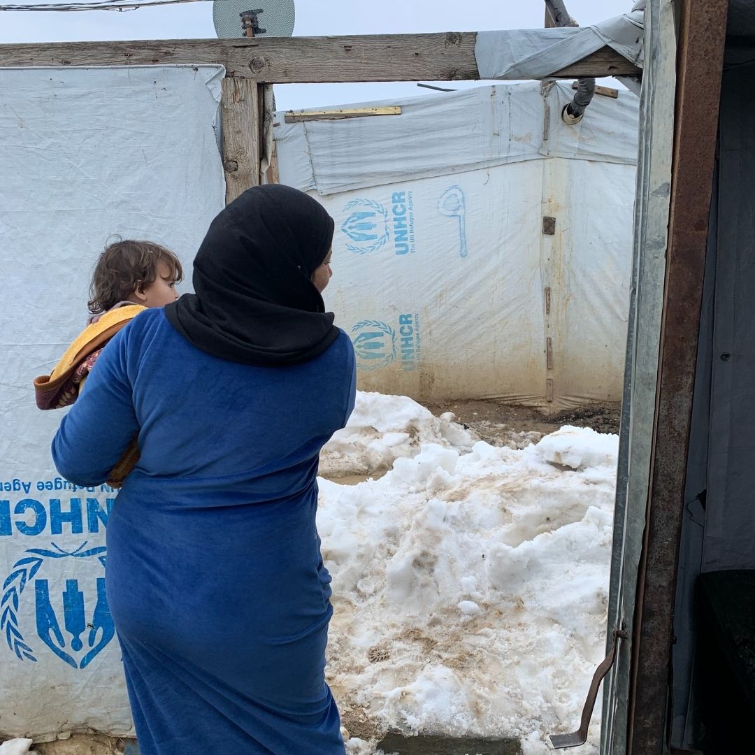 A Syrian woman, holding a small child is standing outside a canvas shelter looking at the mound of snow in front of her.
