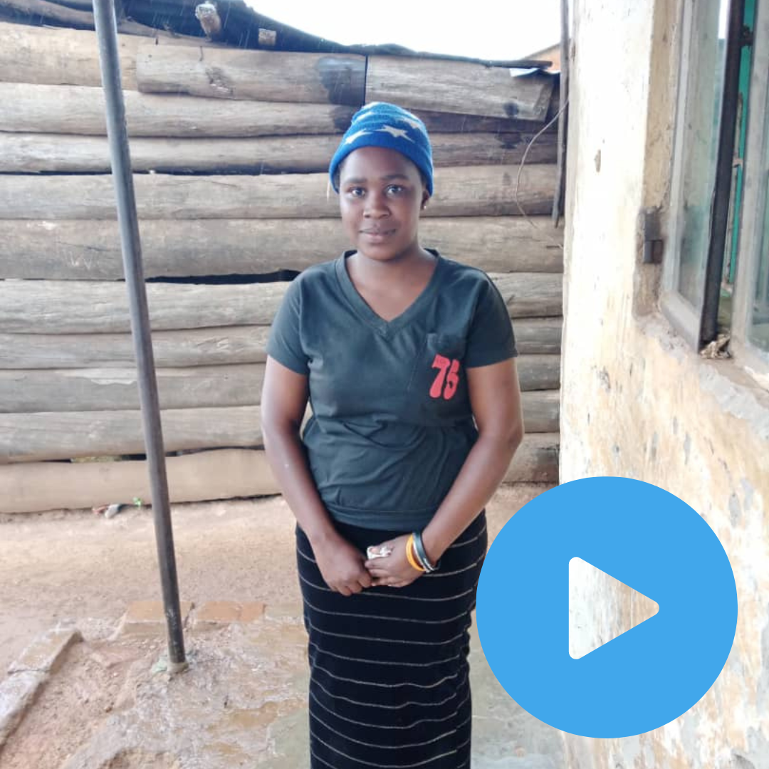 16 year old Leila in Uganda, is stood outside her home in Jinja wearing a blue bandana and grey tshirt and a stripy long black skirt.