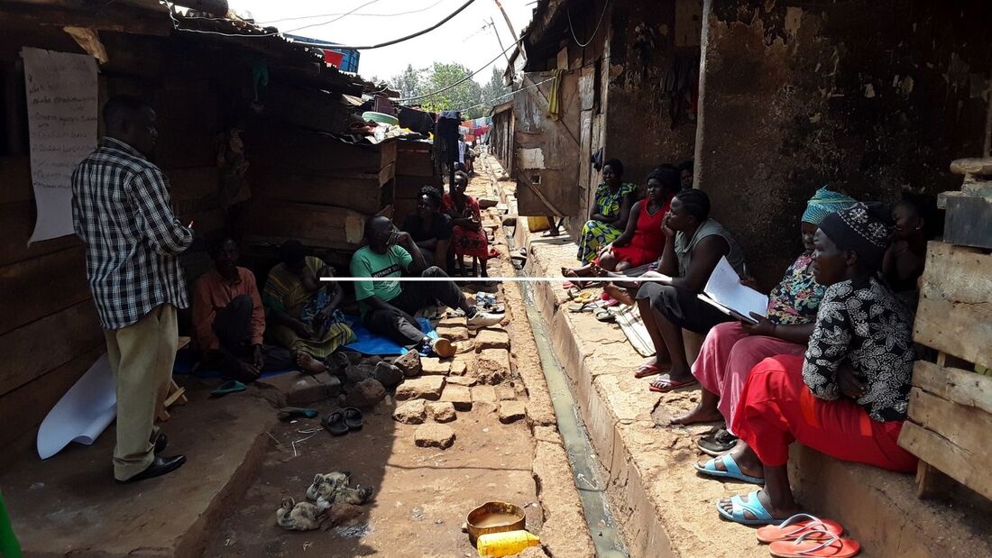 Along a narrow slum street in Loco, Uganda, men and women are gathered, sat under the shade of the buildings to listen to a workshop led by a Ugandan man wearing a checked shirt
