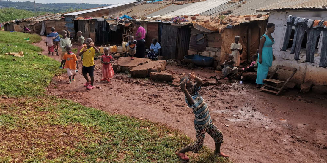 A slum community in Uganda with a line of makeshift houses made from corrugated metal and tarpauline sheets. They are lined up along a dirt road with patches of grass on the opposite side of the track. There are around 11 children walking and playing outside the houses. One small boy closest to the camera is waving. Three adults are standing around the children. 