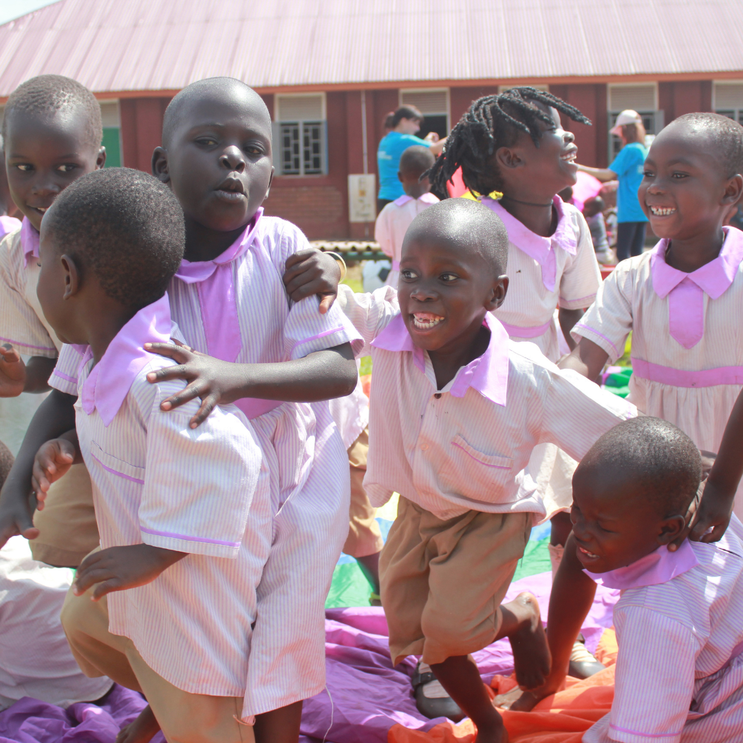 Image shows 7 young Ugandan children aged 3-4 playing outside their school building. They are wearing purple uniform are all smiling and laughing as they play together. You can click on the image to read the blog post