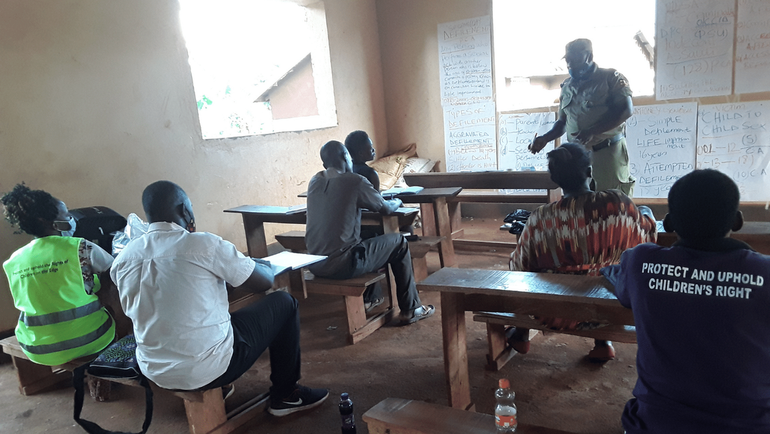 6 members of the Child Protection Team in Mafubira taking part in a workshop in a small classroom, led by a police officer. 