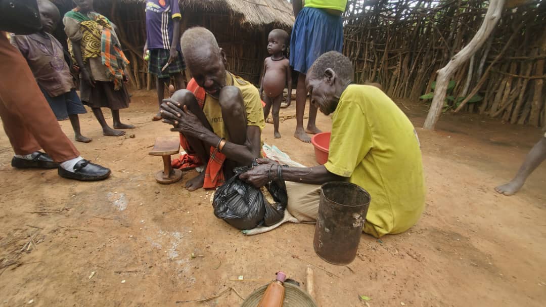 Two older Ugandan men are crouched on the floor wearing yellow shirts holding a black plastic bag full of maize