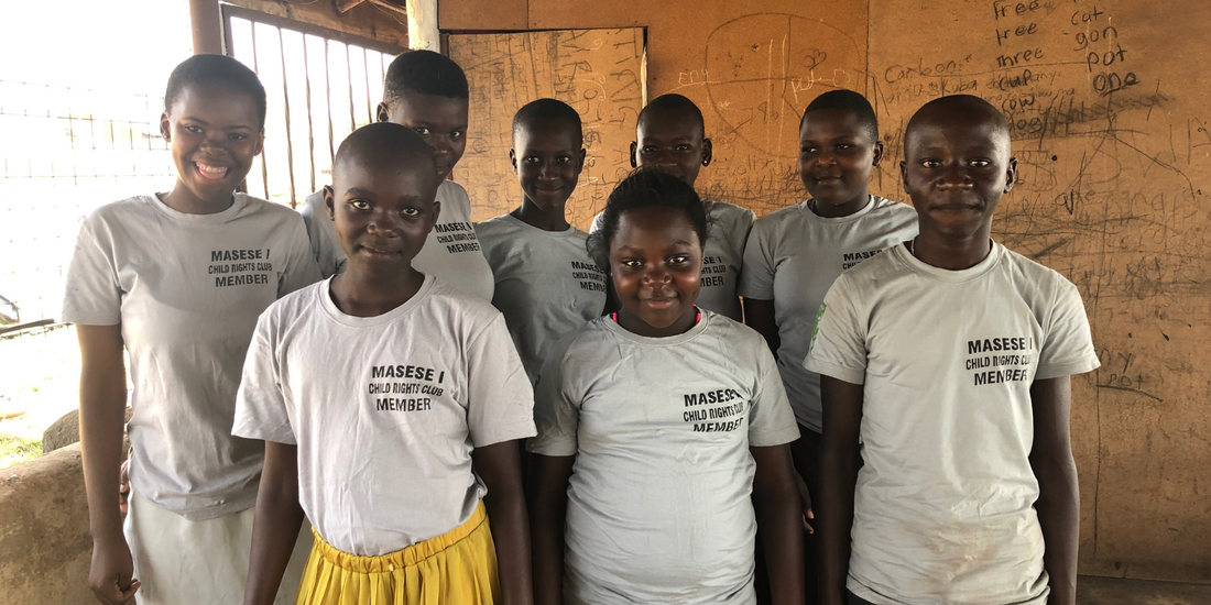 8 Ugandan young teens standing in two rows wearing white tshirts with 'Masese I Child Rights Club member' on. They are standing in what appears to be a classroom next to the window. They are looking at the camera and smiling.