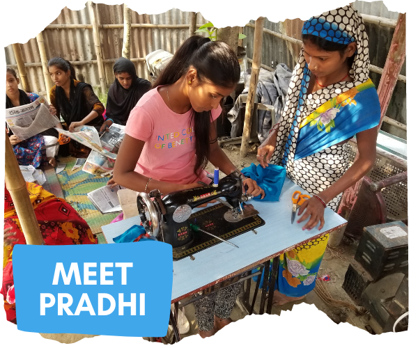 Pradhi sat at an old style sewing machine in a classroom made from bamboo sticks. A teacher is looking over her as she sews a blue piece of fabric. Pradhi is looking at the machine and wearing a pink tshirt. Three other girls are sat on the floor behind her reading newspapers. 