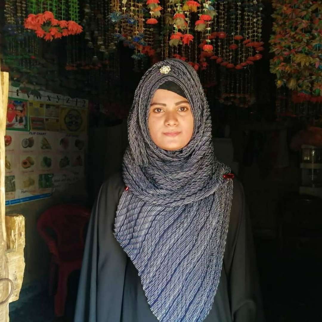 Rishma, a teacher in the Kutupalong camp. She is stood in one of the classrooms in the camp and is waring a glue and white patterned headscarf. She is looking directly at the camera with a neutral expression. You can click this photo to read more of her story