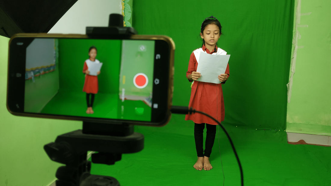 A young girl from Bangladesh is stood in a 'green room' wearing her school uniform and bare feet. She is reading from a script in front of a phone recording her on a stand. 