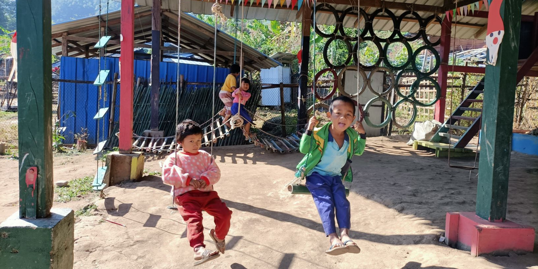 Four young Kachin children playing in the playground outside their ECD Cent in Kachin state. In the front are a young girl and boy sat on two swings made from bamboo and rope. The girl is wearing a pink jumper and red bottoms, she has short dark hair and is looking at the camera, not smiling. The young boy is wearing a bri swings made from bamboo and rope. The girl is wearing a pink jumper and red bottoms, she has short dark hair and is looking at the camera, not smiling. The young boy is wearing a bright green jacket and blue trousers and is sticking is tongue out, looking happy. The play are is undercover, but outside, the sun is shining. In the background there is a bamboo play bridge with two young girls climbing across it. There is another climbing area made of old bike tyres hanging from the roof of the shelter. In the background there are trees and another basic building made from wood with a metal roof.ght green jacket and blue trousers and is sticking is tongue out, looking happy. The play are is undercover, but outside, the sun is shining. In the background there is a bamboo play bridge with two young girls climbing across it. There is another climbing area made of old bike tyres hanging from the roof of the shelter. In the background there are trees and another basic building made from wood with a metal roof.