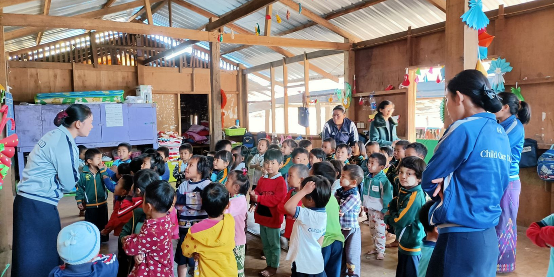 A large classroom made up of a timber frame and metal roof. Inside the classroom are colourful decorations and a large group of around 20 young Kachin children. They are looking at a female teacher in front of them. There are four other adults in the classroom. The children all look happy and focused on what the teacher is saying. 
