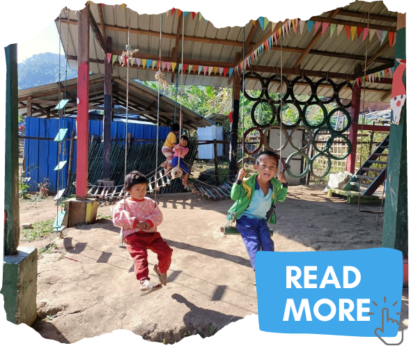 Two young displaced children from Kachin State, Myanmar. They are sat on swings in one of the centres that we support. There are tyre climbing frames behind them and coloured bunting. Attached button image invites you to click to read more. 
