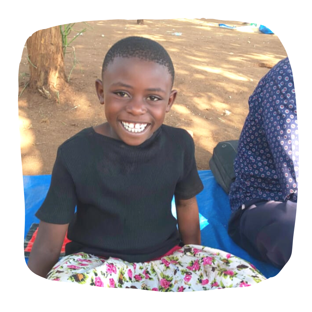 Sifa, a young Congolese girl sat on a blue mat under a tree wearing a black tshirt, floral skirt and smiling.