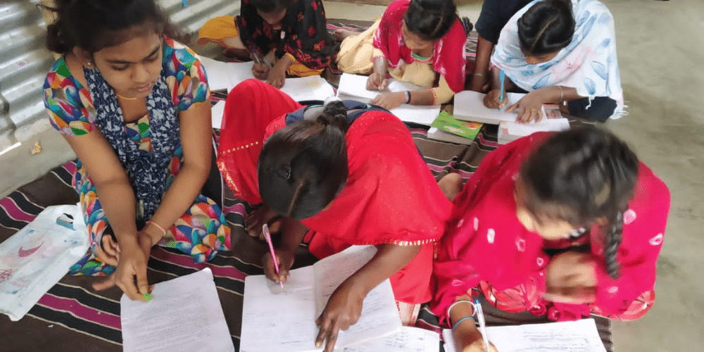 6 young Indian women working in A4 workbooks, writing. You cannot see their faces, but they appear to be concentrating on their work. They are sat in two rows of three. Three are wearing bright coloured Sari's. 